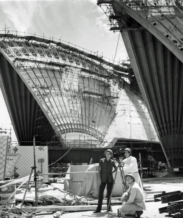 Sails of the Sydney Opera House under construction. A camera crew of three men pose amidst the construction site.