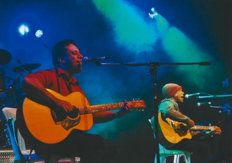 Musician Dave Adern with Archie Roach on stage singing.