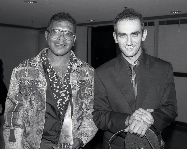 A black and white photograph of Archie Roach and Paul Kelly. Archie holds an ARIA award in his hand. They are both smiling.