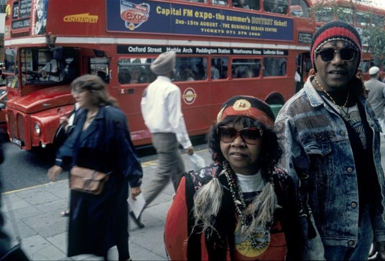 A photograph of Archie Roach and Ruby Hunter standing on a busy street in London. People walk behind them and there is a red double decker bus in the background. 