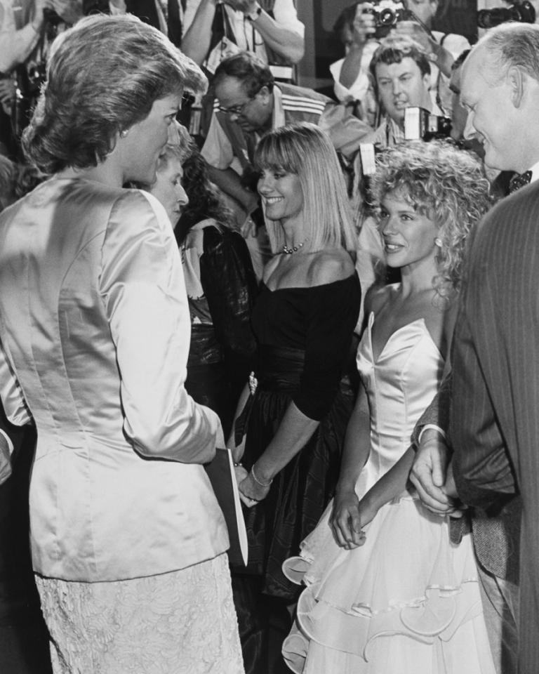 Kylie Minogue, Diana Princess of Wales and Olivia Newton-John stand next to one another in this black and white photograph. Kylie is awestruck looking up at Diana who is much taller.