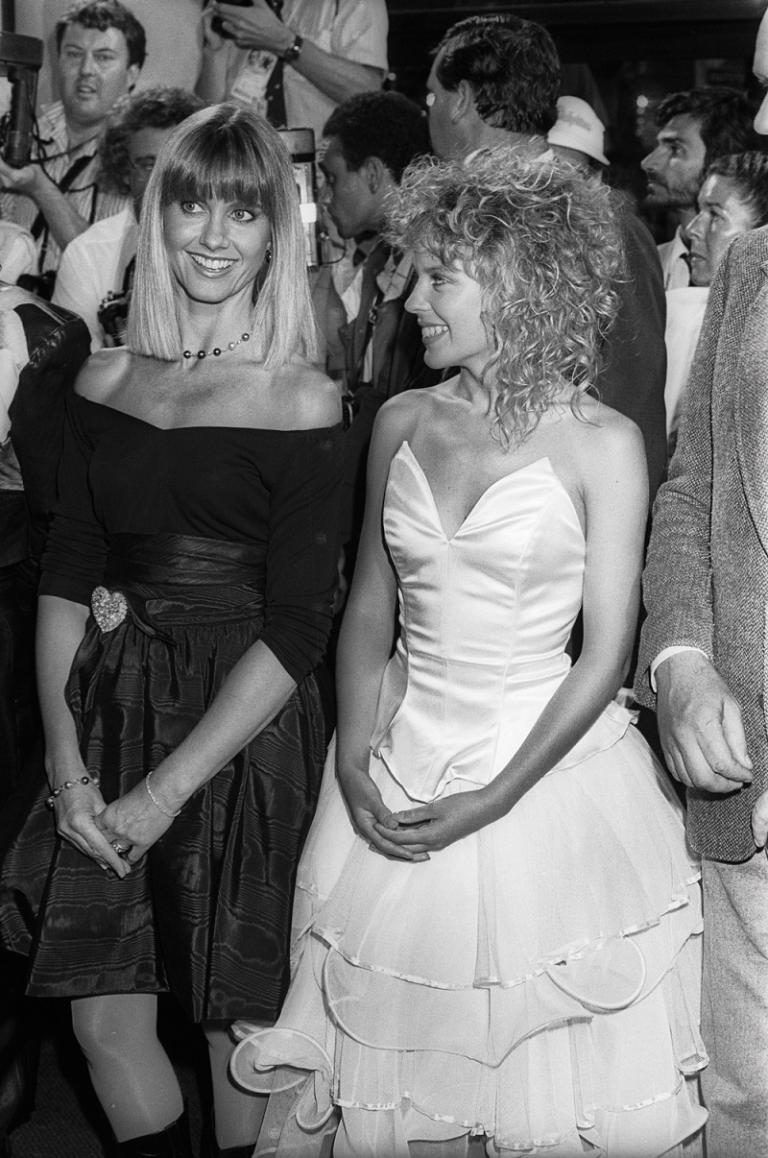 Kylie Minogue and Olivia Newton-John stand next to each other in this black and white photograph. Kylie looks at Olivia adoringly.