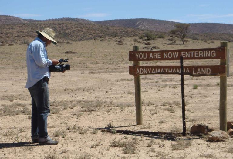 A man holding a video camera standing in the outback in front a sign that says 'You are Now Entering Adnyamathanha Land'