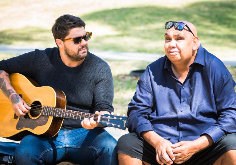 Indigenous singer/songwriter Dan Sultan, pictured outdoors seated with a guitar next to TV presenter and performer Kutcha Edwards.