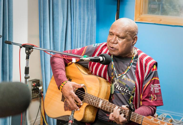 Indigenous singer/songwriter Archie Roach, pictured seated with a guitar in front of a microphone stand