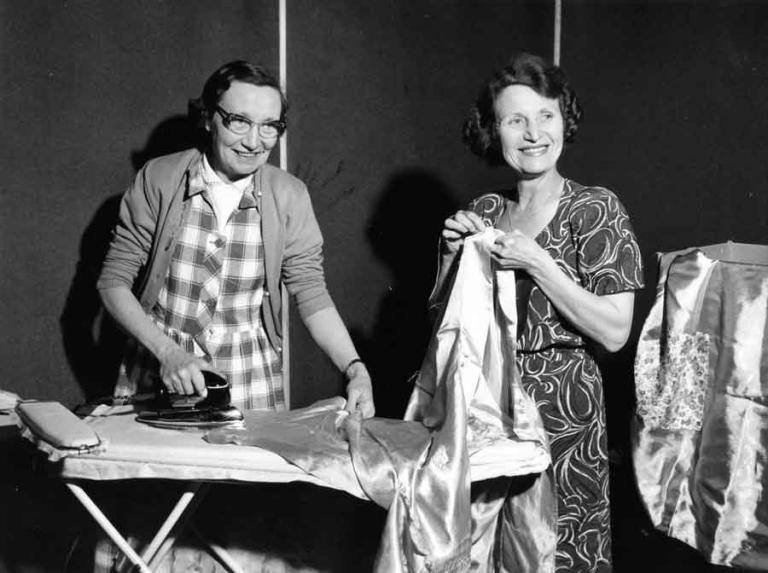 Two women are smiling for the camera. One of them is ironing a piece of clothing on an ironing board and the other is holding some fabric.