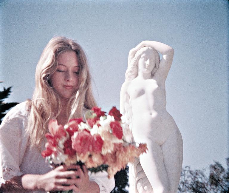 Miranda (Anne Lambert) holding flowers next to a statue in the grounds of the College