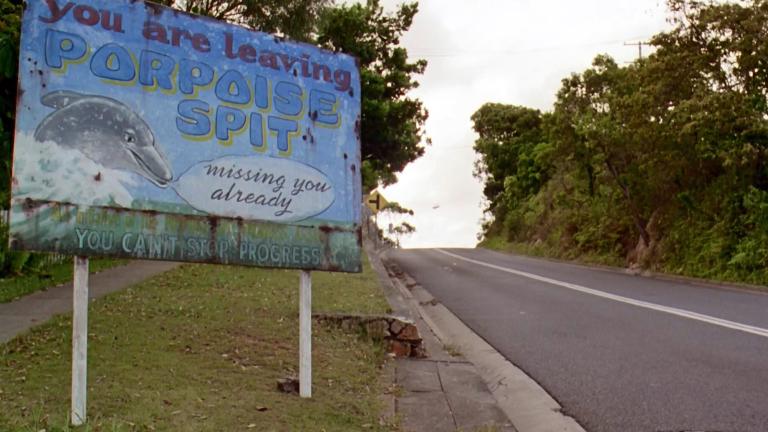 Sign on the side of the road at the end of 'Muriel's Wedding' reads "You are leaving Porpoise Spit". A picture of a dolphin with a speech bubble next to it says "missing you already" and the words "You Can't Stop Progress" are written at the bottom.