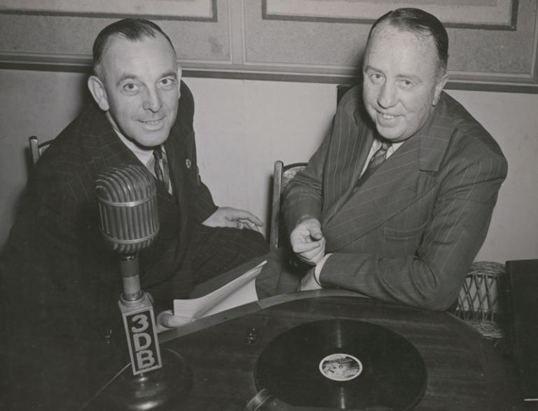 Two men are looking up at the camera. They had a record and a microphone in front of them. The microphone says 3DB.
