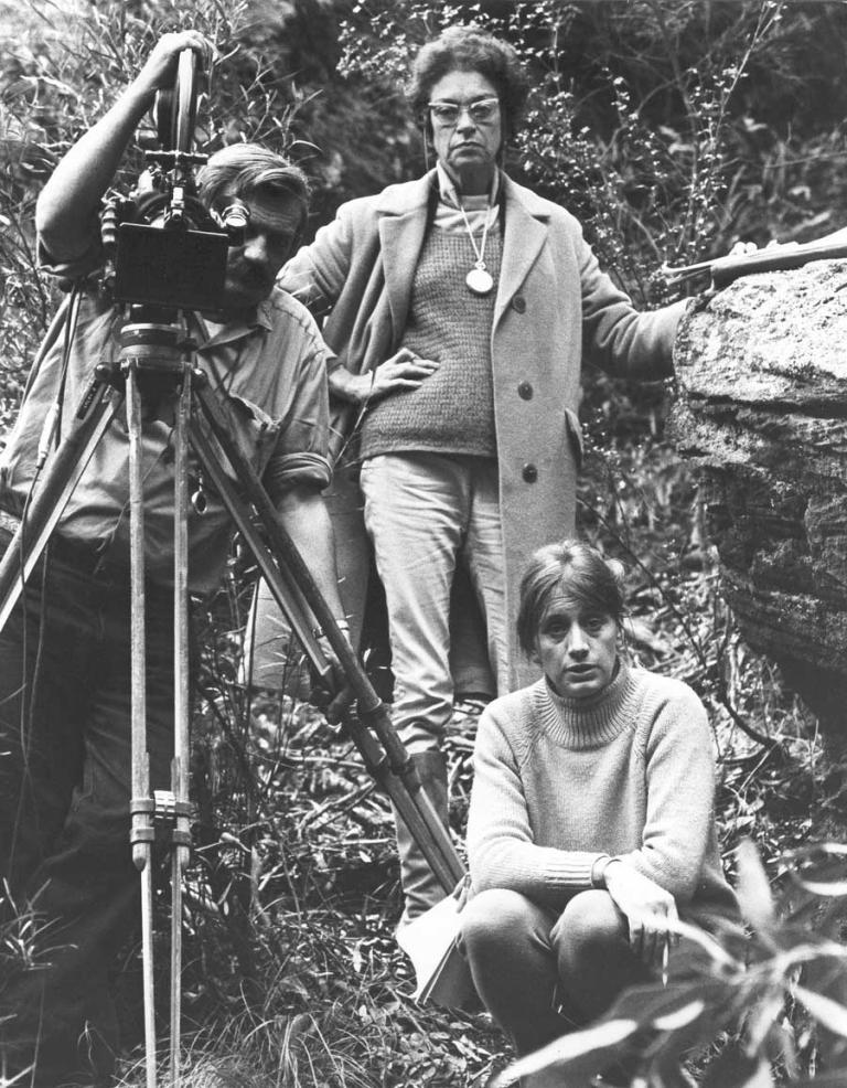 A portrait of three people. A man is holding a film camera on a tripod. One woman is standing with her hand on her hip and the other woman is crouching down with papers on her lap. They are in a natural setting.