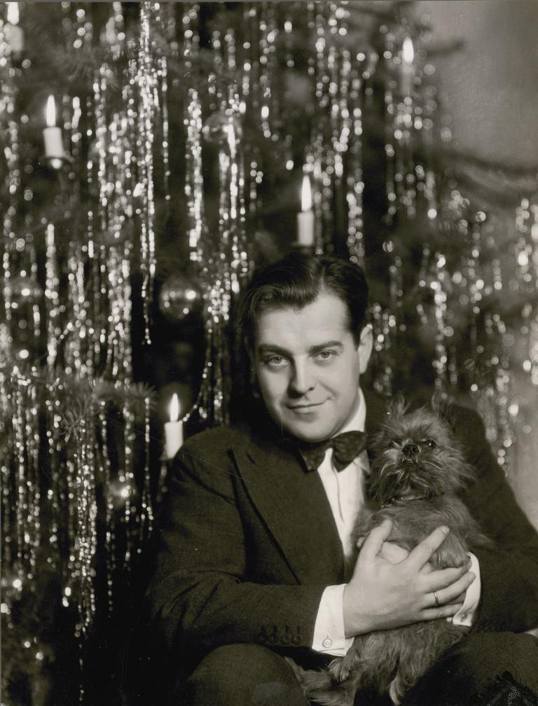 Hermann Speelmans, wearing a suit and holding a small dog, sitting in front of a Christmas tree.