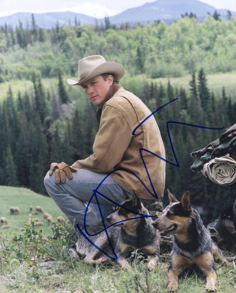 Heath Ledger in a hat and jacket as farm hand Ennis Del Mar from the film Brokeback Mountain. There are mountains and rolling hills and trees in the background.