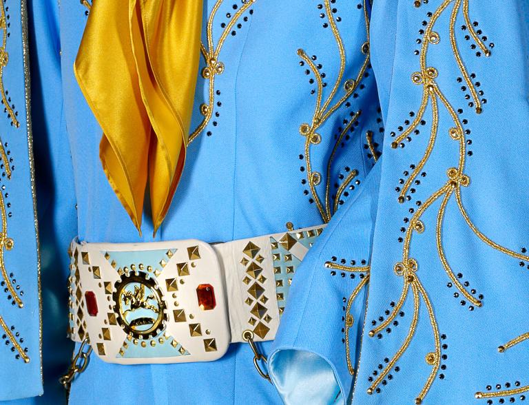 Close up on part of a costume from the film Elvis detailing gold embroidery on a blue fabric, with a yellow scarf and large belt buckle featuring rhinestones.
