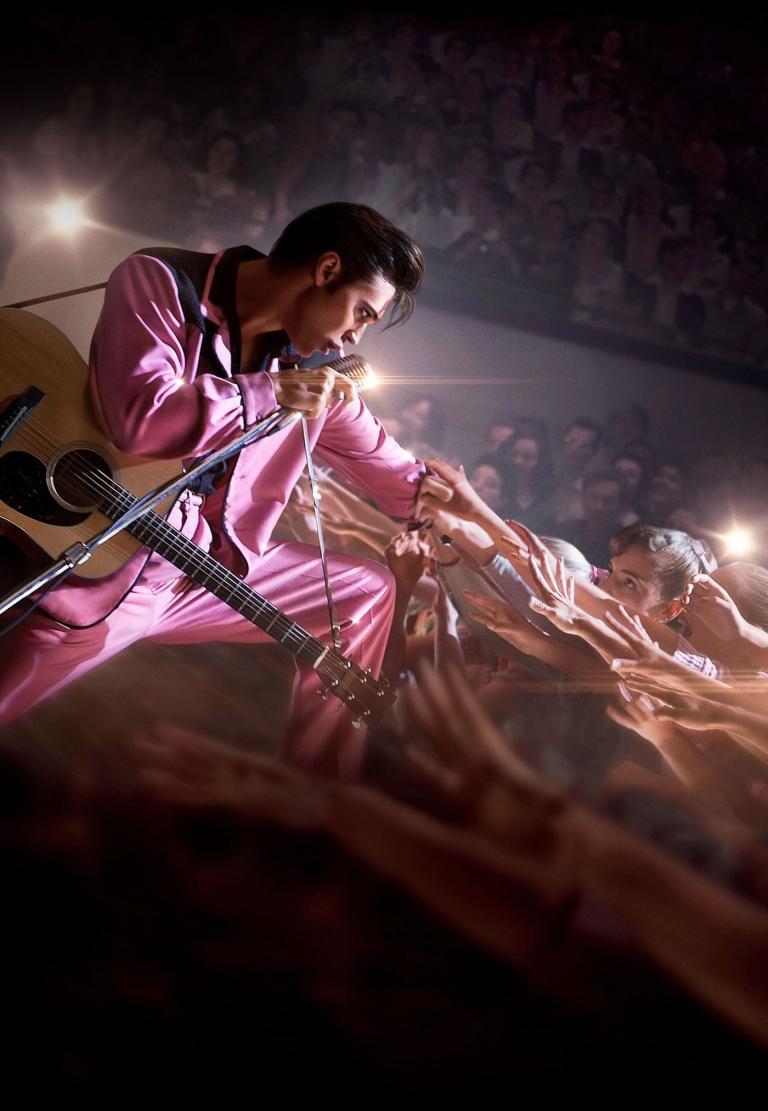 Actor Austin Butler dressed as Elvis Presley in a pink and black suit pictured on a stage. He is holding a microphone with a guitar strapped over his shoulder. He is leaning forward into the audience who are all reaching out towards him.