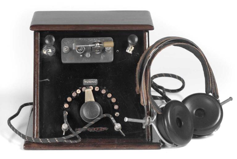 A radio and headphones dated 1925. The radio is in a wooden box with a dial on the front and the headphones are attached with a cord and have leather ear pads and trim.