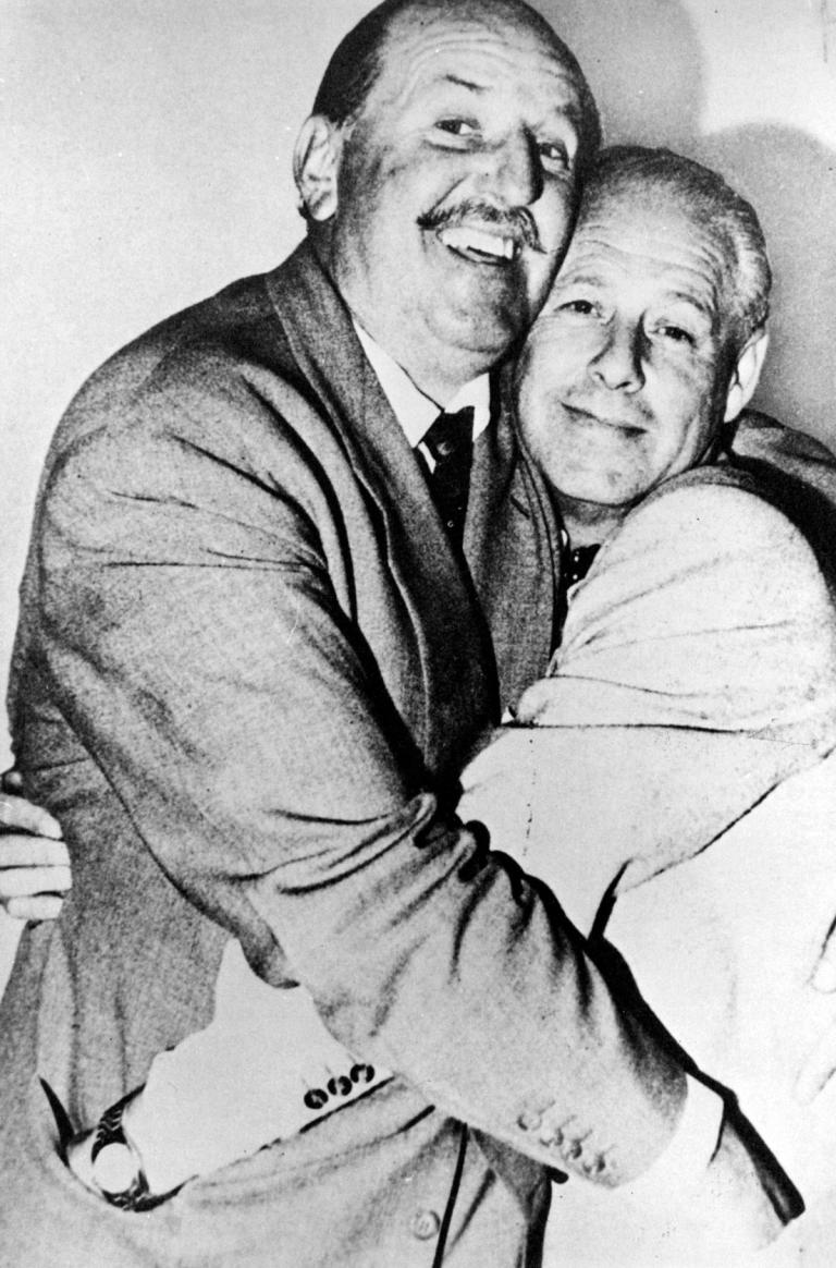 Two middle-aged men embracing and smiling. They are looking at the camera and have their hands in each other's pockets.