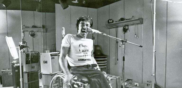 Singer Jeff St John in a recording studio, seated in a wheelchair, wearing headphones and singing with eyes closed into a microphone.