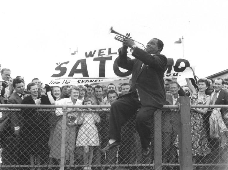 Louis Armstrong at Sydney airport in 1956, He's sitting on the top rail of a wire fence, playing the trumple to a crow gathered there to welcome him.