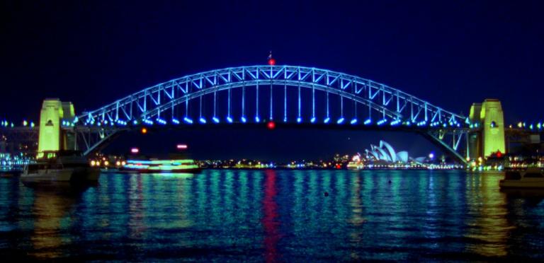A nightime photo of the Sydney Harbour Bridge showing it all lit up. The bridge arch is blue with red lights at the top and bottom centre. The pylons are lit yellow. The water shows the reflections of the passing ferry and the Sydney Opera House.