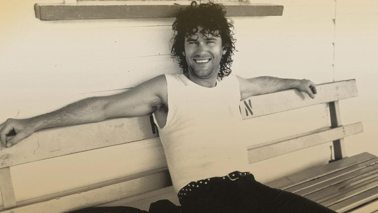 Jimmy Barnes seated on a bench, wearing a white singlet with arms outstretched and smiling at camera.