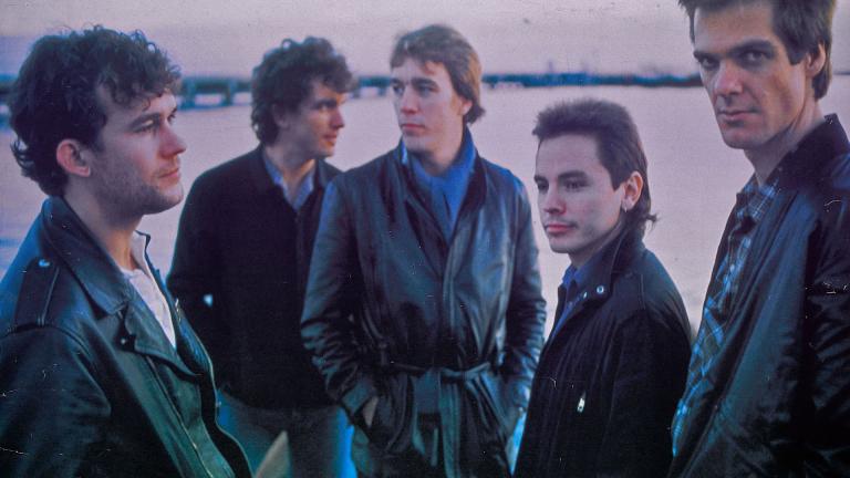 Five band members of Cold Chisel standing on a beach with a pier in the background. They are dressed in leather jackets and looking away from camera. 