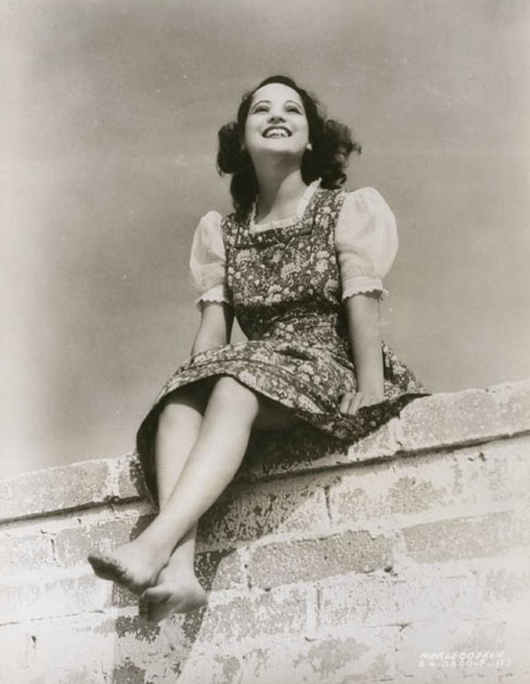Publicity shot from below of Merle Oberon sitting on a wall, bare foot and wearing a floral dress.
