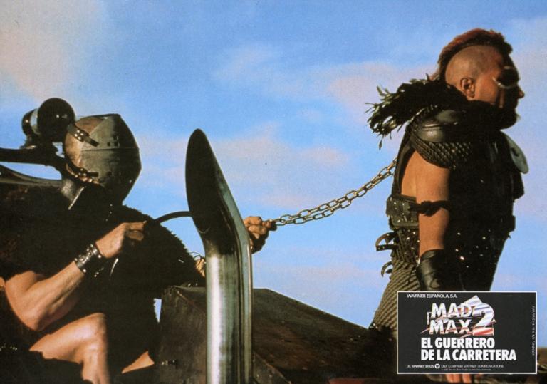 Mad Max 2 lobby card showing Wez (Vernon Wells) chained by the neck by Humungus (Njell Nilsson). 