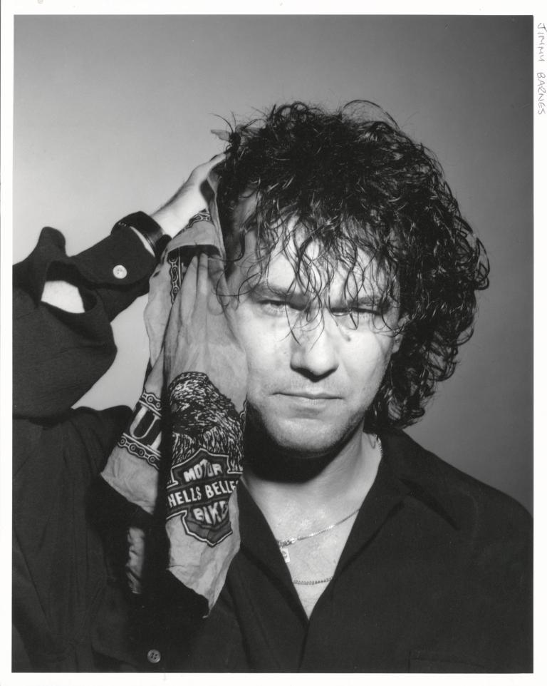 Head and shoulders shot of Jimmy Barnes wearing a black shirt and looking at camera. He is holding a bandana with a motorcycle club logo on it next to the side of his head. 