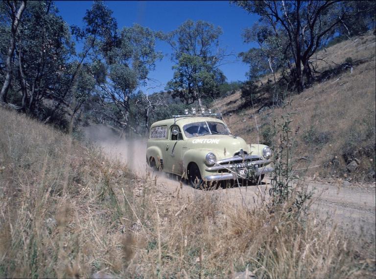 Film still of a cream cvoloured FJ Holden panel van racing along a dusty road.
