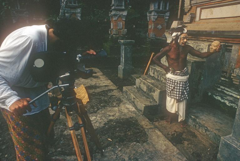 A camera man filming a balinese man in cultural dress and mask