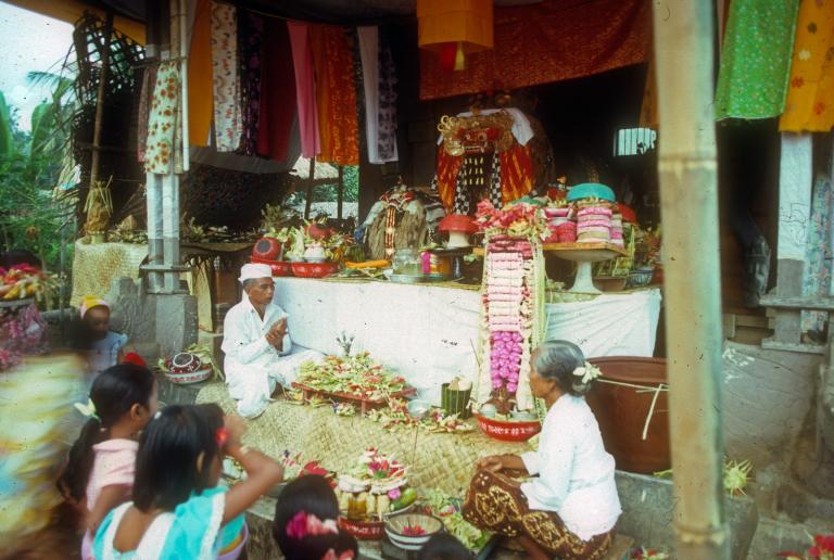 A Balinese ceremony showing a group of people seated in front of what looks to be a small temple with many bright, colourful offerings at the front.