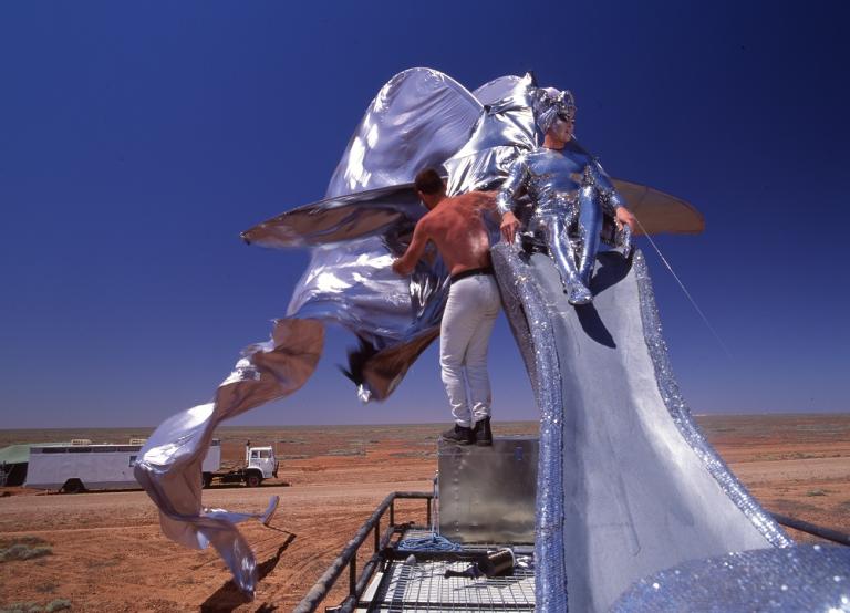 Guy Pearce sitting on a giant silver stilleto shoe on top of the Priscilla bus.