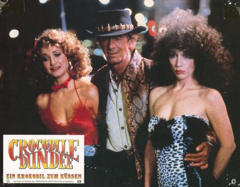 Lobby card depicting Paul Hogan as Mick Dundee with his arms around two New York City prostitutes