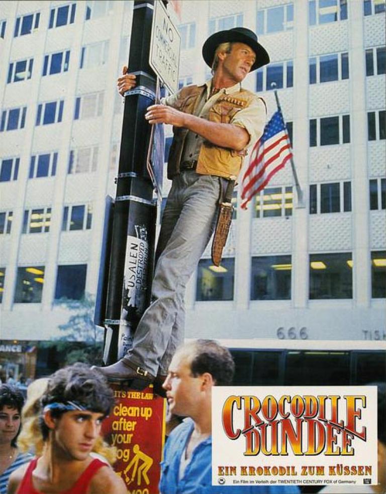 Lobby card depicting Mick Dundee standing up high, hanging on a light pole in New York City