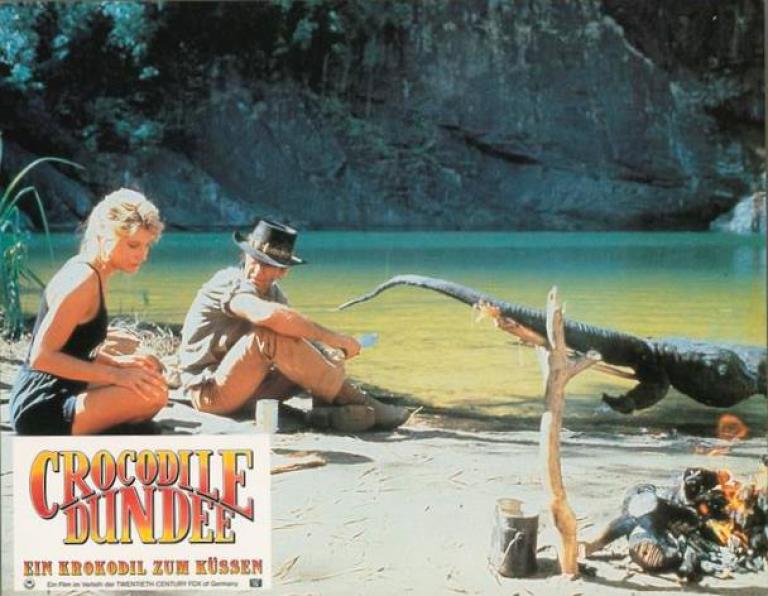 Lobby card depicting Linda Kozlowski as Sue Charlton and Paul Hogan's Mick 'Crocodile’ Dundee in the outback with a crocodile roasting on a spit made of tree branches