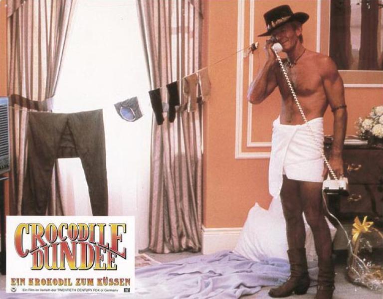 Lobby card depicting Mick Dundee talking on the phone in his hotel room while he stands wrapped in a towel. He has made a makeshift laundry for his clothes across the window.