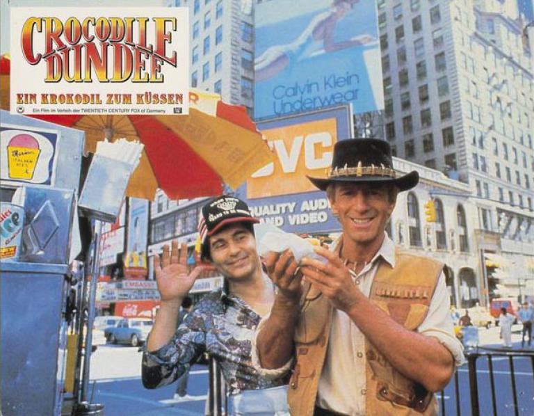 Lobby card depicting Paul Hogan as Mick Dundee holding a hot dog and smiling for the camera in New York City