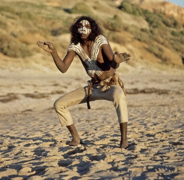 David Gulpilil as Fingerbone Bill in a production still from Storm Boy. He is striking a dance pose standing on a beach and painted with white paint on his face and torso