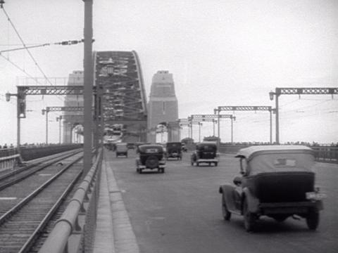 Black and white still of 1930s cars travelling North over the Sydney Harbour Bridge. The railway line is on the left next to the cars.