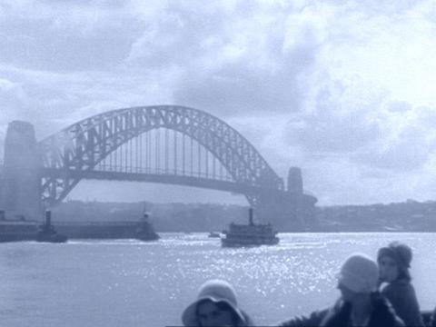A blue tinted black and white still of the Sydney Harbour Bridge as seen from a ferry. Clouds and mist give the bridge a magical aura.