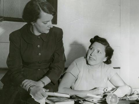 One woman sitting at a desk and the other sitting on the edge of the desk.