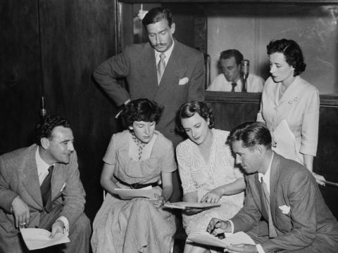 Radio actors huddled around looking at a script.