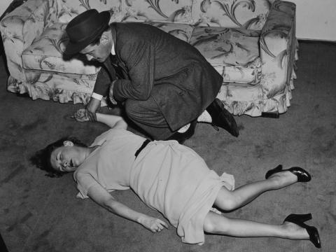 A man in a suit and hat crouches over a murdered woman