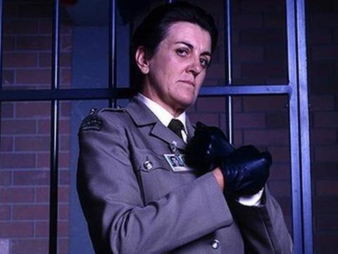 Maggie Kirkpatrick in her role as prison officer Joan Ferguson. She's standing in front of the prison bars, wearing her uniform and her trademark black leather gloves. 