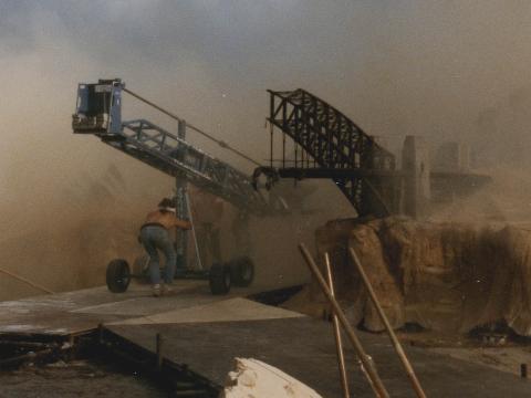 A crew member pushes a crane next to a model set of post-apocalyptic Sydney in some sand dunes. The buildings are about the size of the person. An arch of the destroyed Sydney Harbour Bridge and the Opera House is visible.