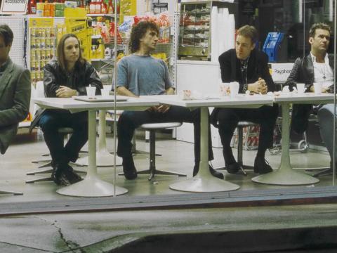 The five members of the band Cold Chisel seated at white plastic tables at a service station. From left to right: Don Walker, Phil Small, Ian Moss, Steve Prestwich and Jimmy Barnes.