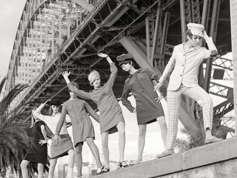 Black and white photo of six female models dressed in fashion from 1964 posing underneath the Sydney Harbour Bridge.