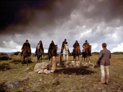 A young man stands with his back to camera looking over a gravesite at the top of a hill. Behind the grave a group of six men on horses look at the young man.