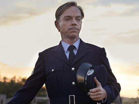 Hugo Weaving as Sergeant Farrat in The Dressmaker. He is standing in uniform alongside a fence.