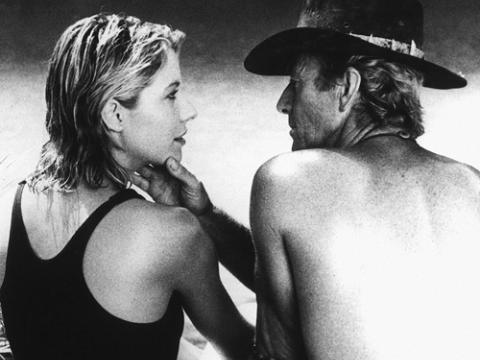 Linda Kozlowski as Sue Charlton and Paul Hogan as Mick ‘Crocodile’ Dundee sitting by a waterhole. He is touching her face and they are staring at each other.